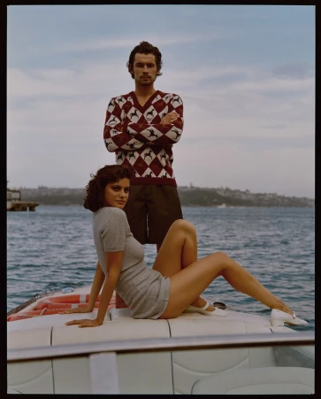 Stylish couple poses on a boat with seascape backdrop. The woman sits in a grey knit dress while the man stands behind in a patterned sweater.
