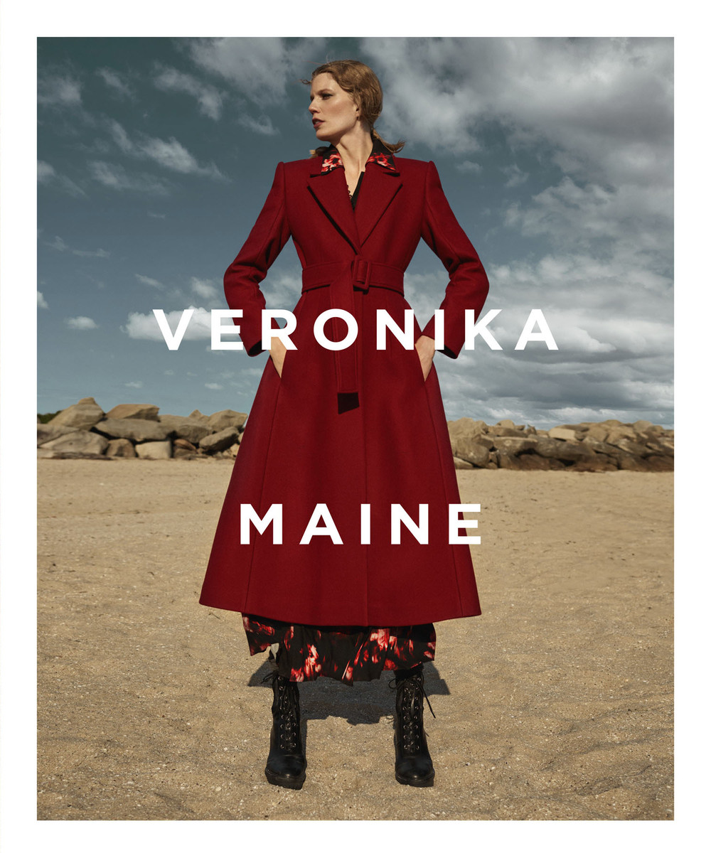Model wearing a long red belted coat standing on a sandy beach under an overcast sky with overlaid white text.