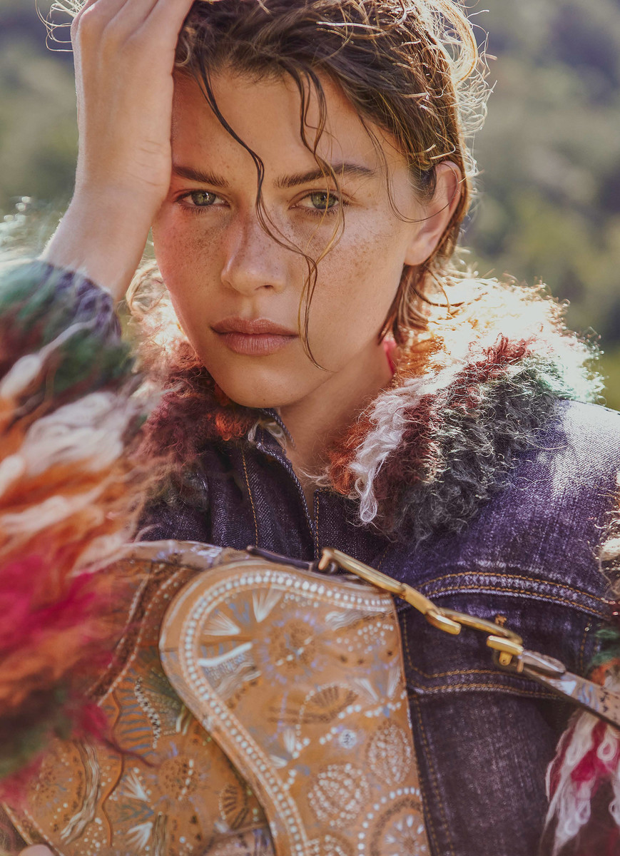 Close-up portrait of a woman with wet hair and freckles wearing a textured denim jacket and holding a patterned tan bag outdoors.