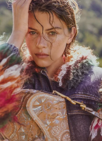 Close-up portrait of a woman with wet hair and freckles wearing a textured denim jacket and holding a patterned tan bag outdoors.