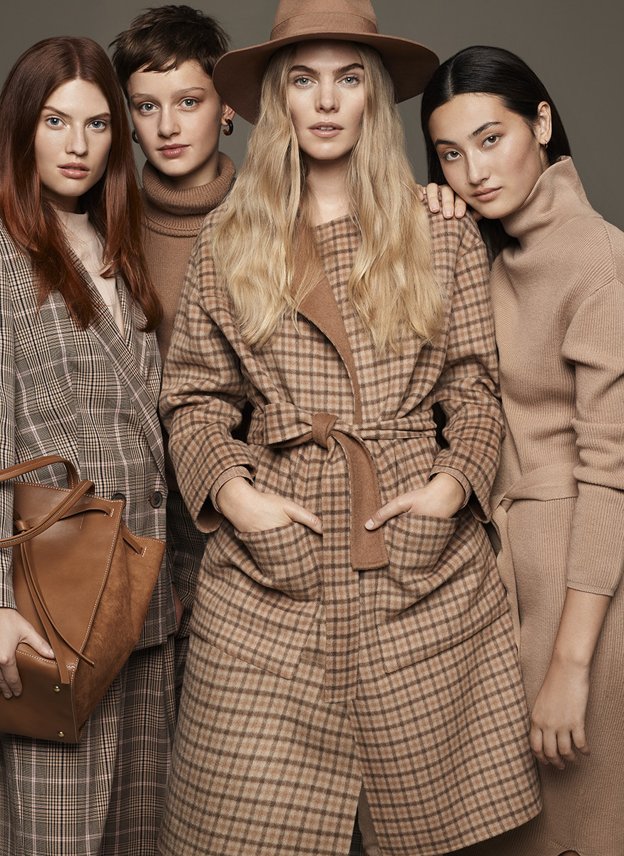A group of four female models in neutral-toned outfits, including a brown plaid coat and hat, pose in a studio setting.
