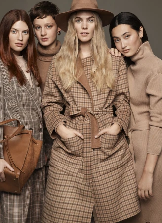 A group of four female models in neutral-toned outfits, including a brown plaid coat and hat, pose in a studio setting.