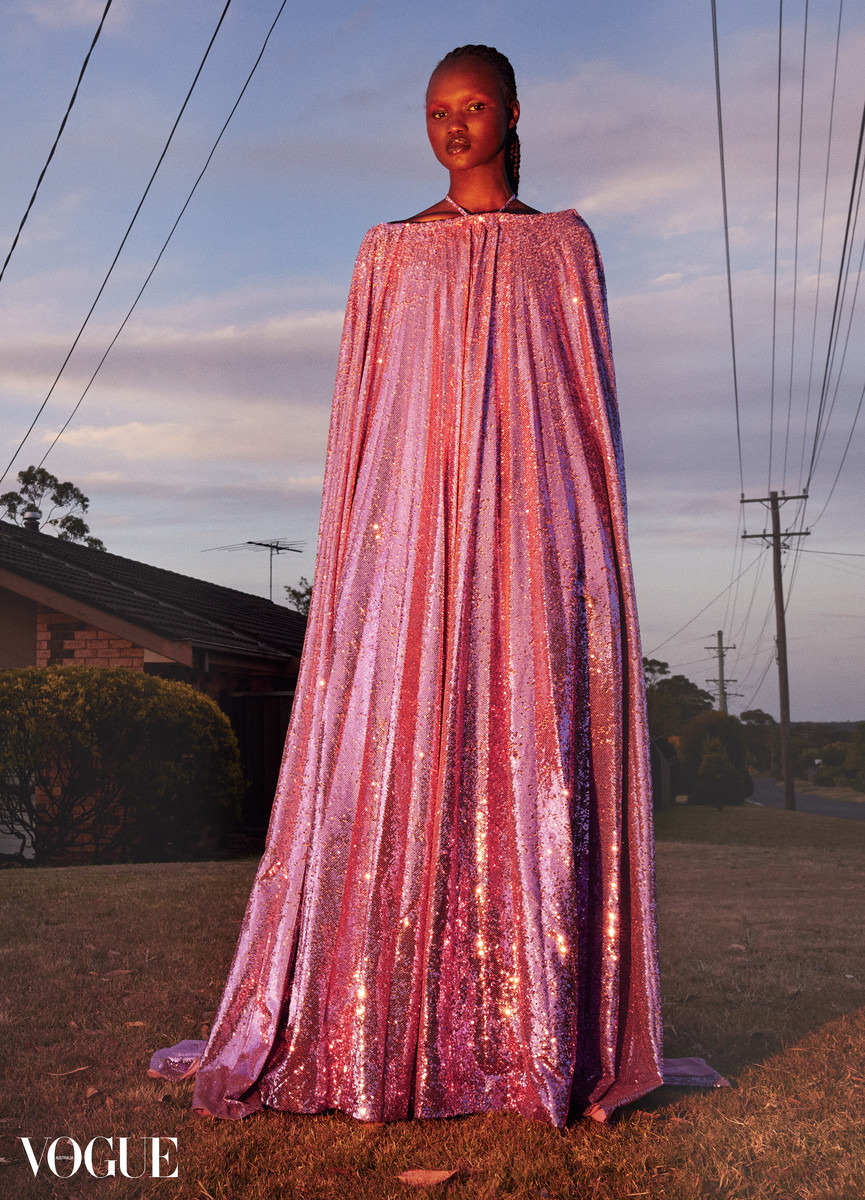 A Black female model poses in a long, pink, shimmering dress and cape in a grassy field with houses and power lines at dusk.