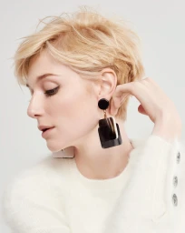 Close-up portrait of a woman with short blonde hair wearing large dark statement earrings and a cream textured sweater against a white backdrop.