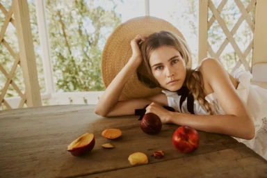 A woman in a wide-brimmed straw hat and white top resting on a wooden table with fresh peaches.