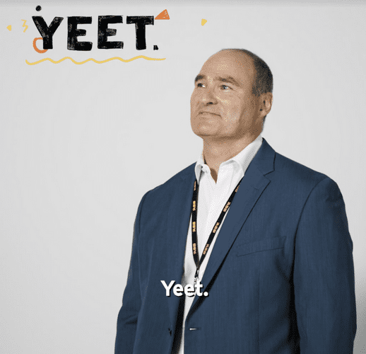 Adult male in a blue suit with a lanyard around his neck, smiling and looking up in a bright indoor setting with text that reads 'Yeet'.