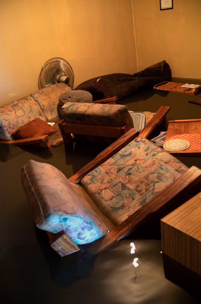 Damaged living room interior with flooded floor, submerged upholstered furniture, and a fan, suggesting a aftermath of flood damage.
