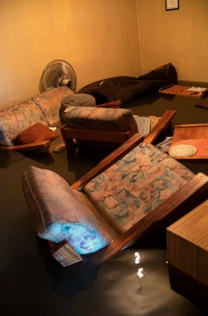 Damaged living room interior with flooded floor, submerged upholstered furniture, and a fan, suggesting a aftermath of flood damage.