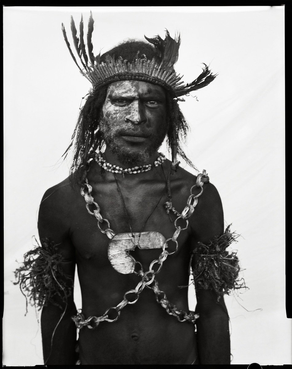 Monochrome portrait of indigenous adult male with tribal paint, feather headdress, and ceremonial necklaces against a plain background.