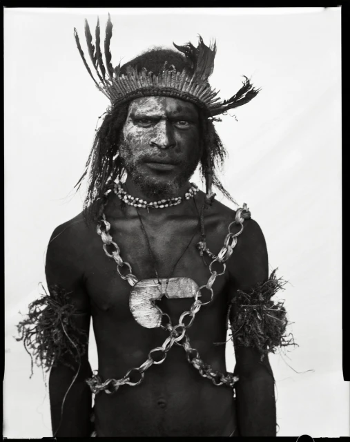 Monochrome portrait of indigenous adult male with tribal paint, feather headdress, and ceremonial necklaces against a plain background.