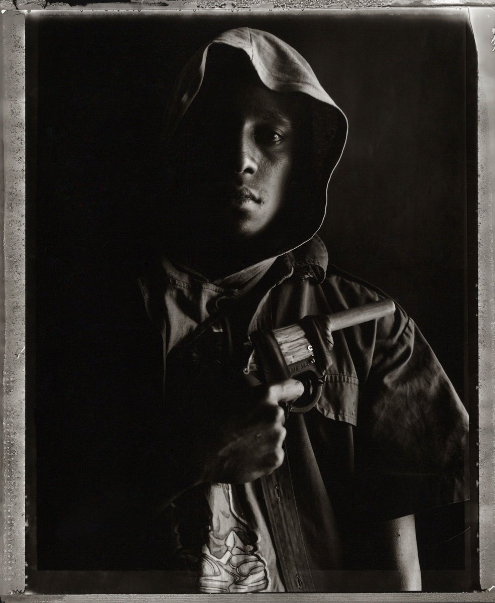Monochrome portrait of a person with dark hair, wearing a hooded jacket, holding an object in their hand, set against a dark backdrop.