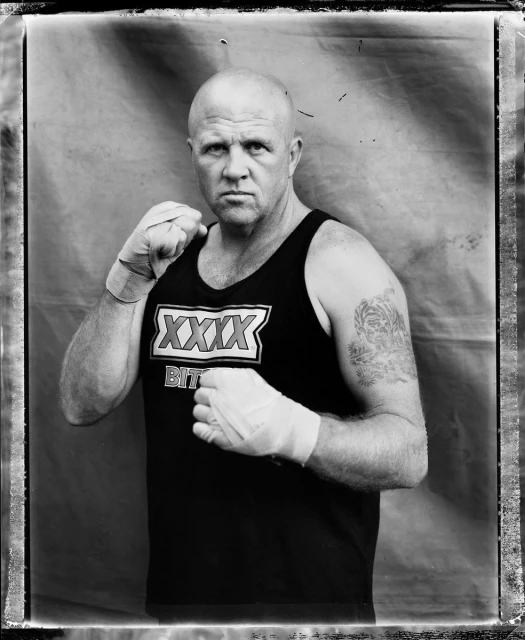 Monochrome portrait of a bald man wearing a XXXX Bitter tank top and boxing wraps, set against a mottled backdrop, framed in a vintage-style border.
