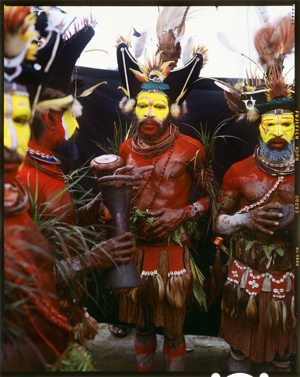 A group of people with painted faces and traditional headdresses and costumes are gathered outdoors, one holding a drum, under natural light.