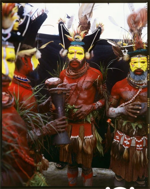 A group of people with painted faces and traditional headdresses and costumes are gathered outdoors, one holding a drum, under natural light.