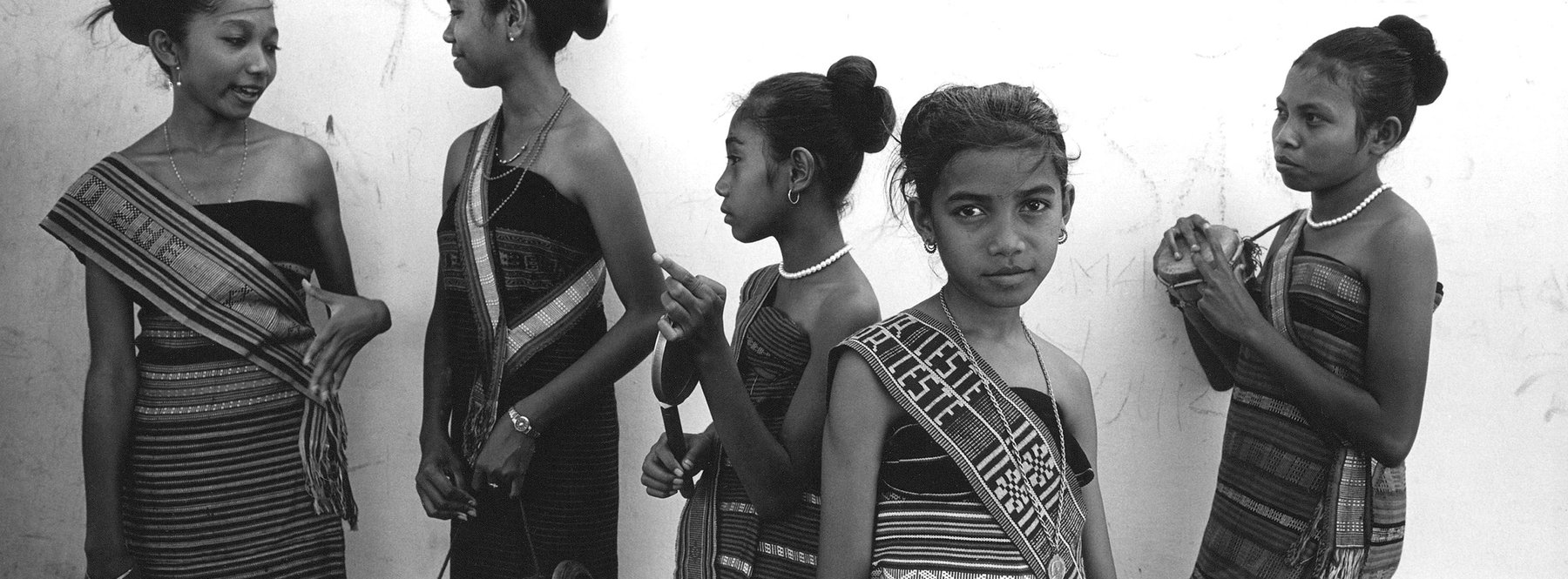 Five young girls in traditional woven clothing stand against a wall in a black and white photograph. One girl holds a small drum.