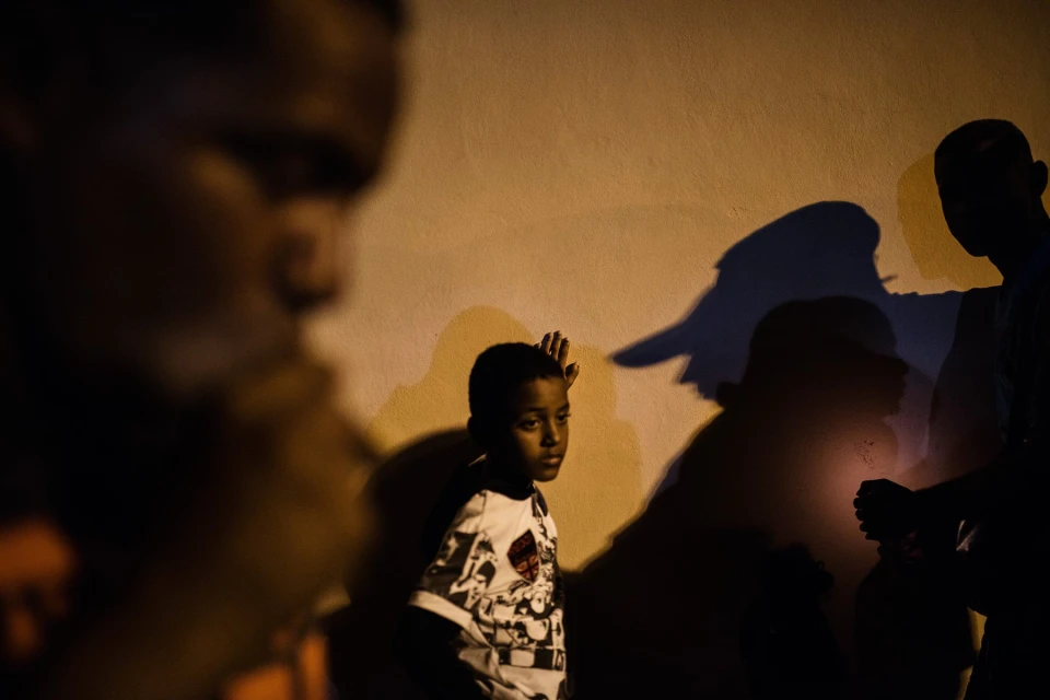 Boy in patterned shirt stands against wall in shadow, with blurred figures and silhouettes suggesting group and nighttime setting.