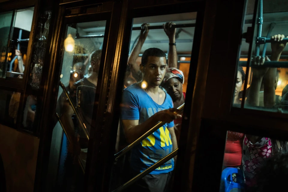 People on a bus at night with a man in a blue shirt in the foreground, a woman wearing a headscarf, and passengers.