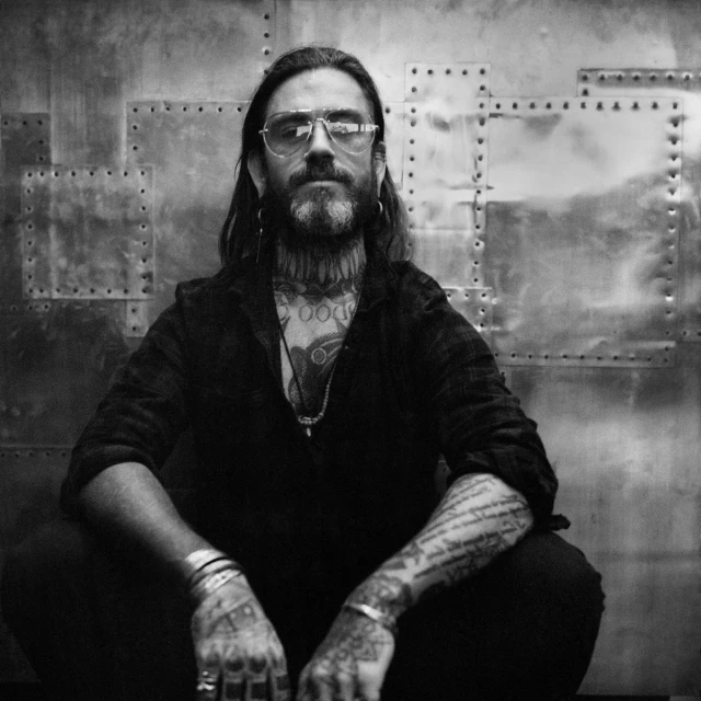Monochrome portrait of a tattooed man with a beard and glasses, wearing a dark shirt, in front of a textured metal background.