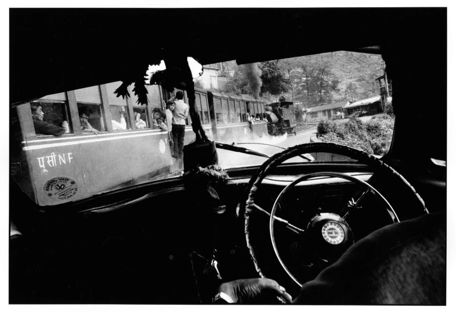 Black-and-white photograph from inside a car looking out at a steam train with people, smoke, and buildings in a mountain setting.