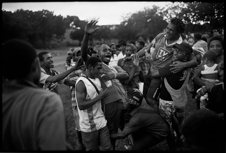Black and white image captures a group of jubilant men celebrating outdoors, one being carried on shoulders amid cheering and excitement.