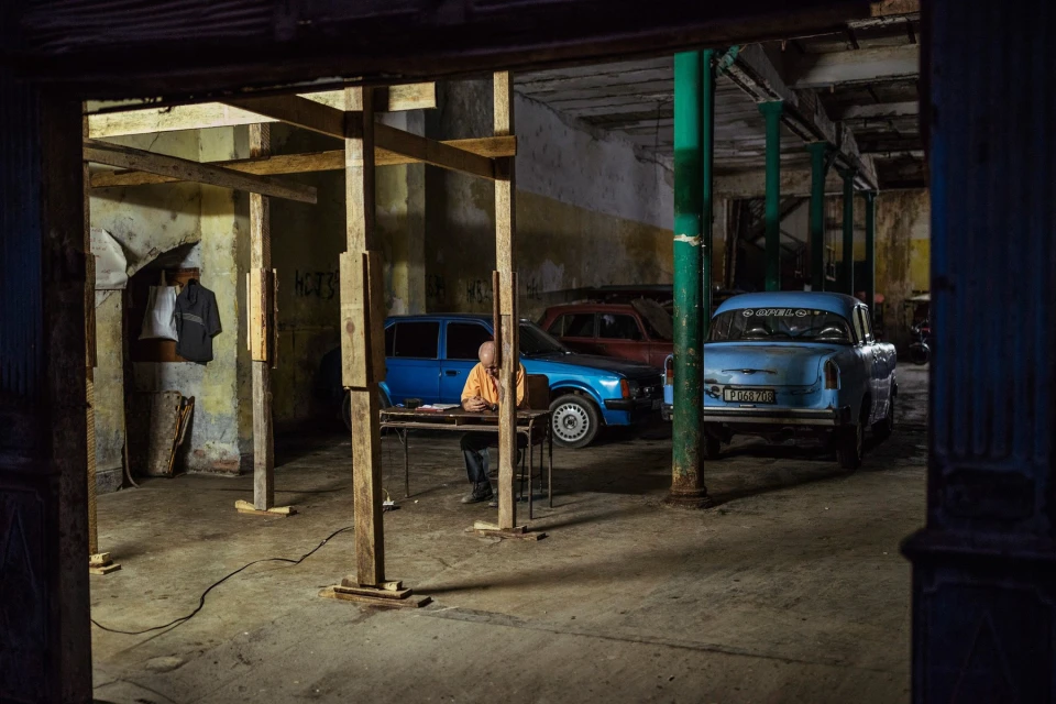 Indoor shot of a man sitting at a table in a large room with old cars, pillars, and wooden scaffolding. Interior view, low light.