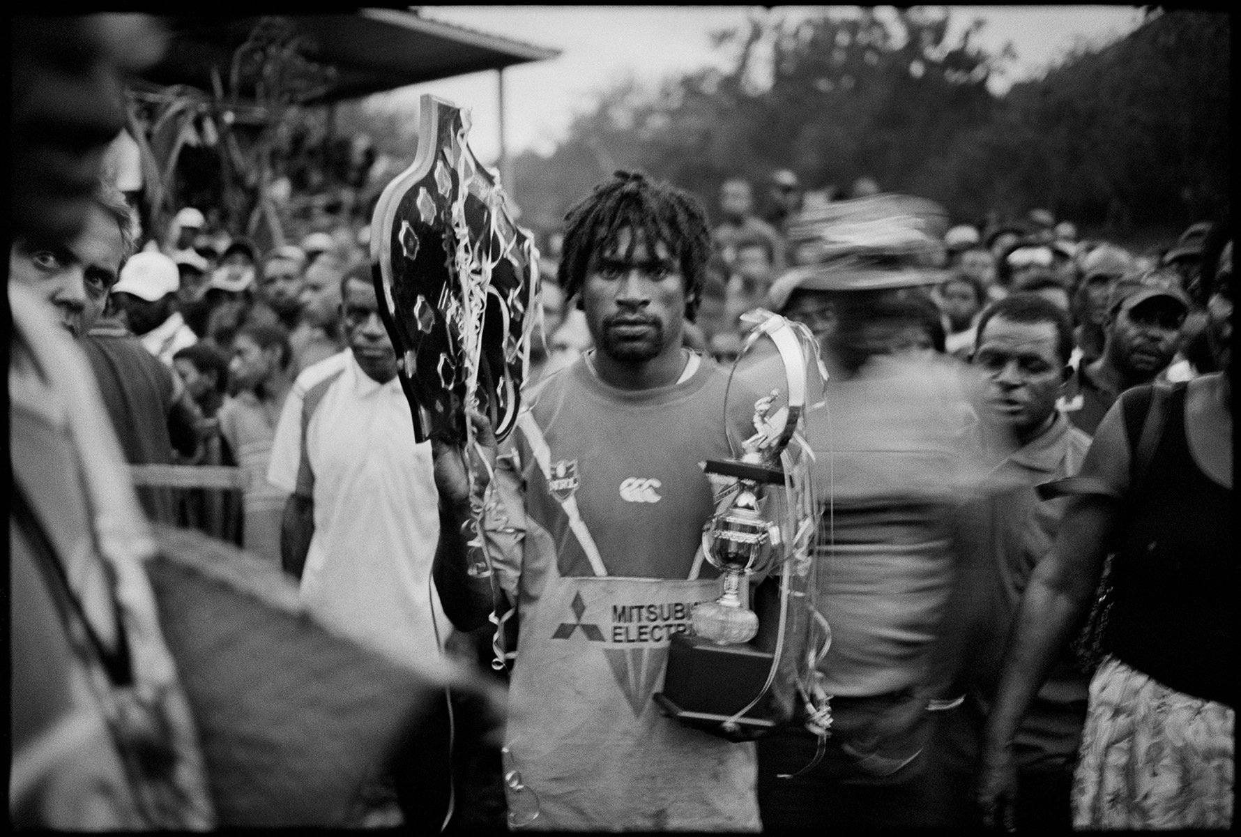 Black and white photo of a man holding trophies in a crowd, wearing a team jersey with the Mitsubishi Electric logo visible.