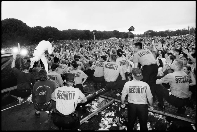 A concert scene captured in black and white, featuring a performer on stage, a crowd, and a security team. The environment suggests an outdoor event.