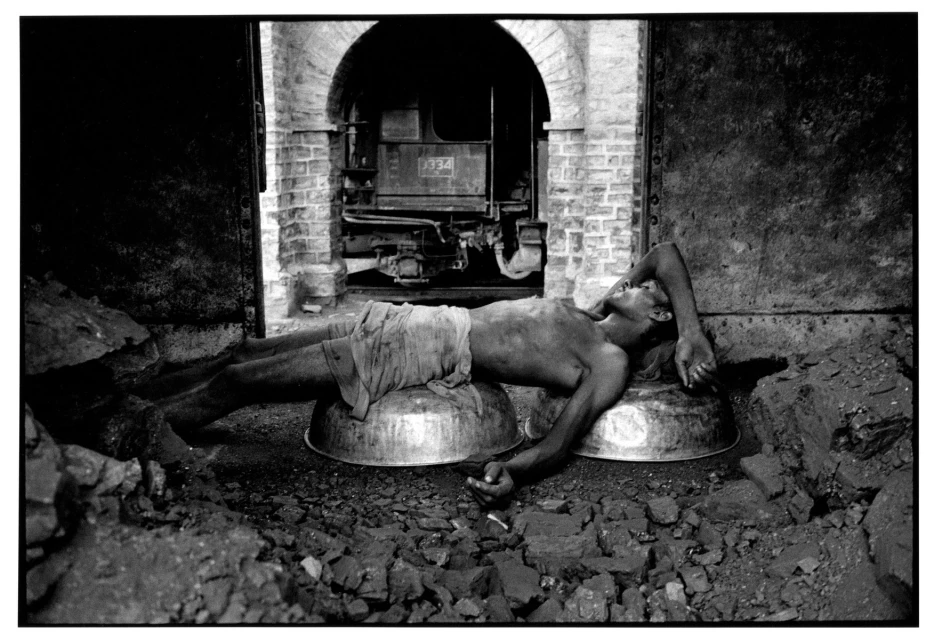 Monochrome image features a man resting on metal basins beside a train in a brick structure, creating a gritty industrial scene.