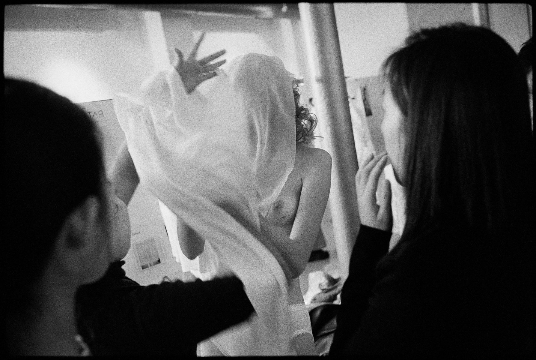 Black and white backstage fashion shoot featuring a model with a sheer white garment, captured in a candid moment with assistants present.