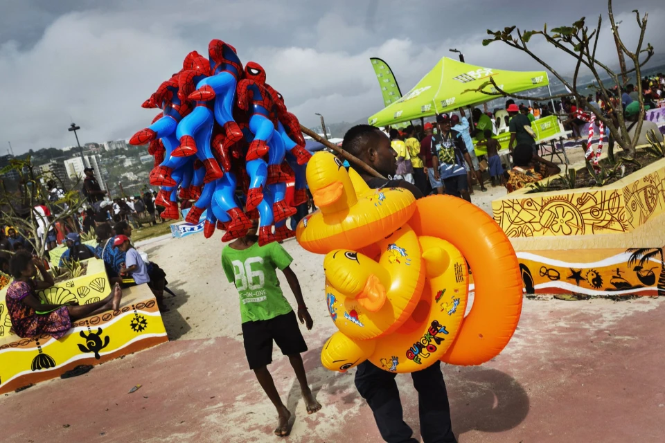Two children walking outdoors, one carrying Spider-Man balloons and the other a stack of duck-shaped inflatable rings, in a public setting.