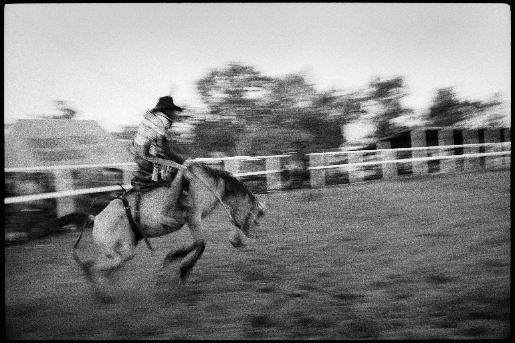 Black and white action shot of a cowboy riding a bucking bronco in an outdoor rodeo arena with motion blur.