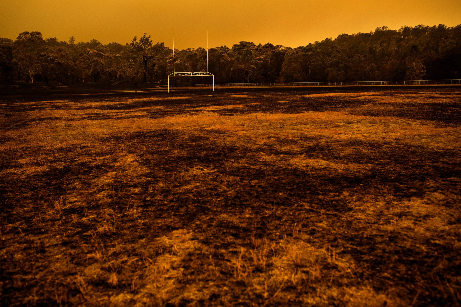 Empty sports field with goalposts, featuring dead grass and trees in the background, bathed in an eerie orange light, wide shot.