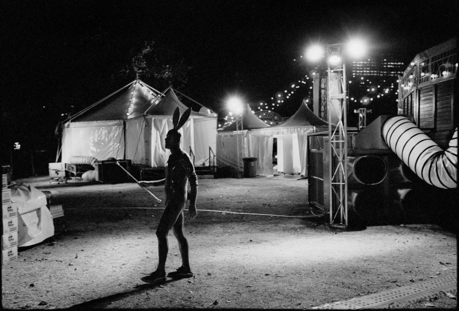 Person in bunny ears walks in front of tents with stage lighting and audio equipment at an outdoor nighttime event.