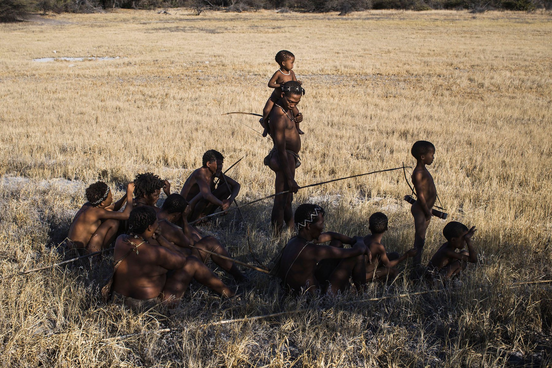 A group of African people are seated and standing in a grassy field, some holding spears in a natural outdoor environment.