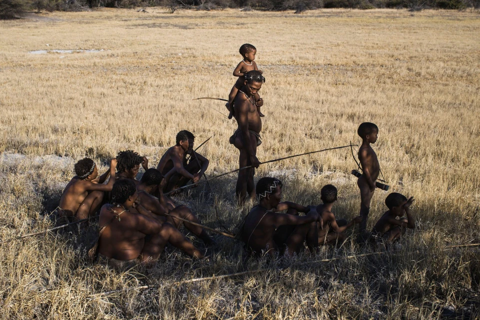 A group of African people are seated and standing in a grassy field, some holding spears in a natural outdoor environment.