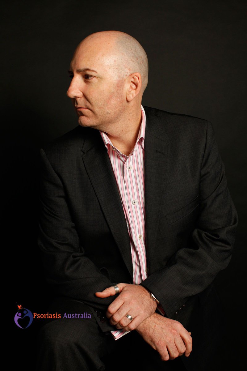 Portrait of a bald man wearing a dark suit and pink shirt, looking to the side against a dark backdrop, showcasing a professional yet approachable demeanor.