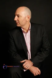 Portrait of a bald man in a dark suit jacket and striped shirt looking away against a dark background