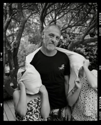 Black and white portrait of a man in a t-shirt outdoors with children holding white fabric.