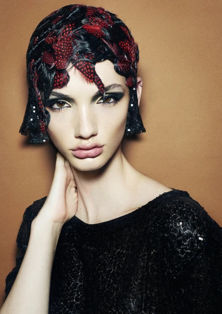 Portrait of a woman with feather and sequin hairpiece, wearing a sequined top against a tan background. Glamour shot for fashion or beauty.