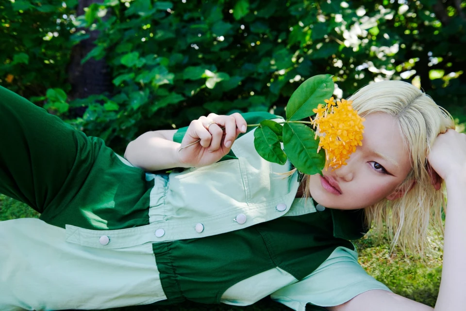A woman with bleached blonde hair and a green patchwork outfit poses in grass with an orange flower held near her face in a close-up shot.