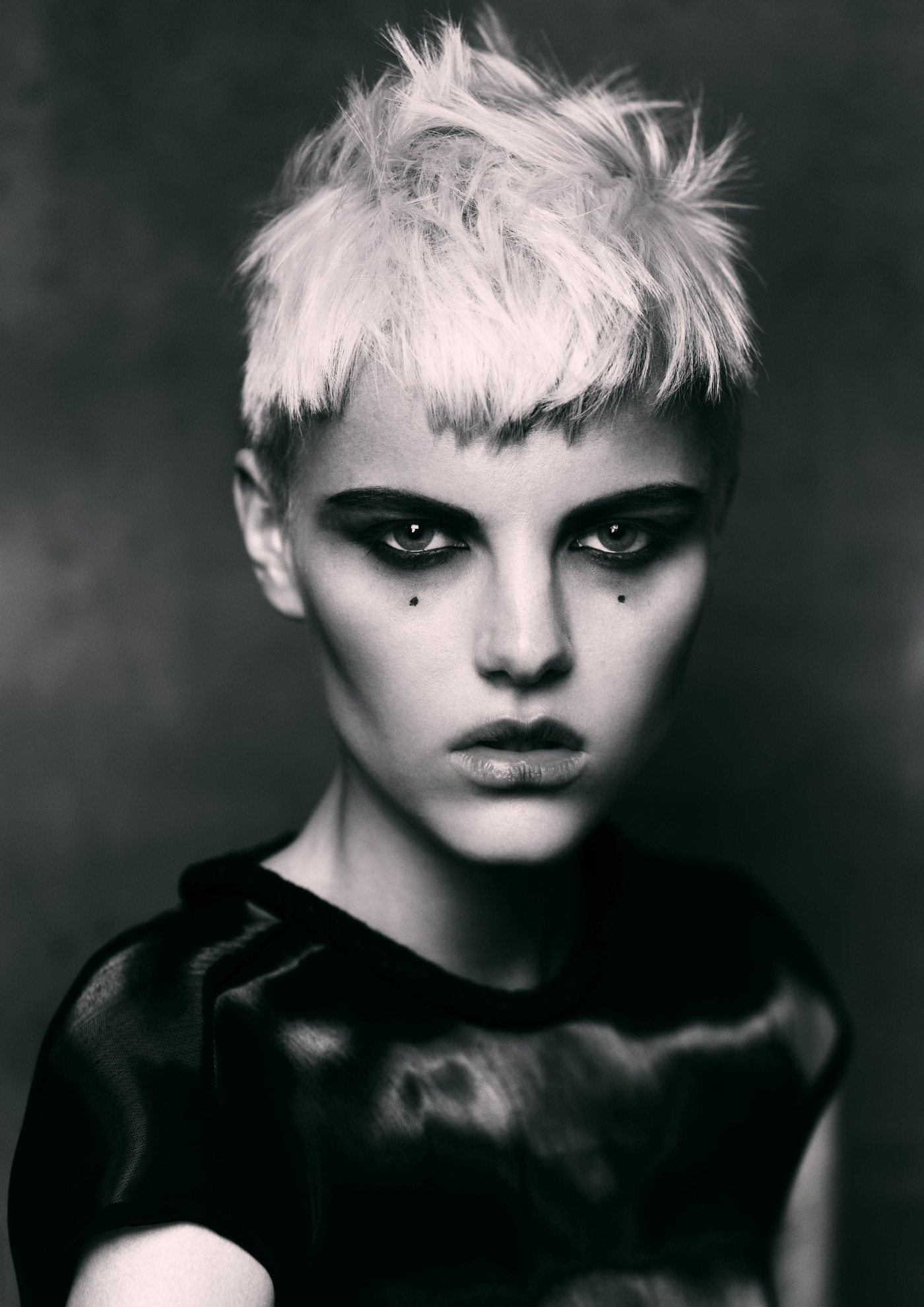 Striking black and white portrait of a woman with short platinum blonde hair, dramatic eye makeup, and a sheer black top against a dark backdrop.
