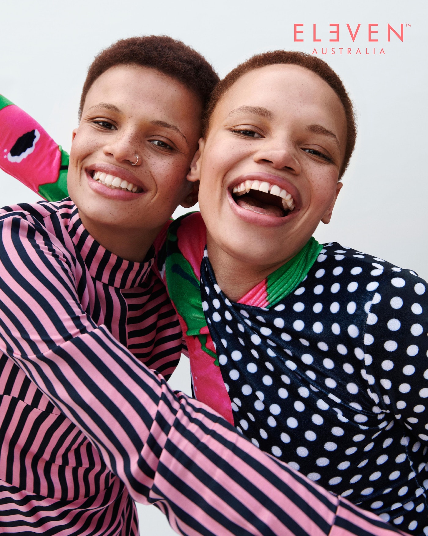 Two smiling women, one in pink stripes and one in black polka dots, hug against a white background in a close-up studio portrait.