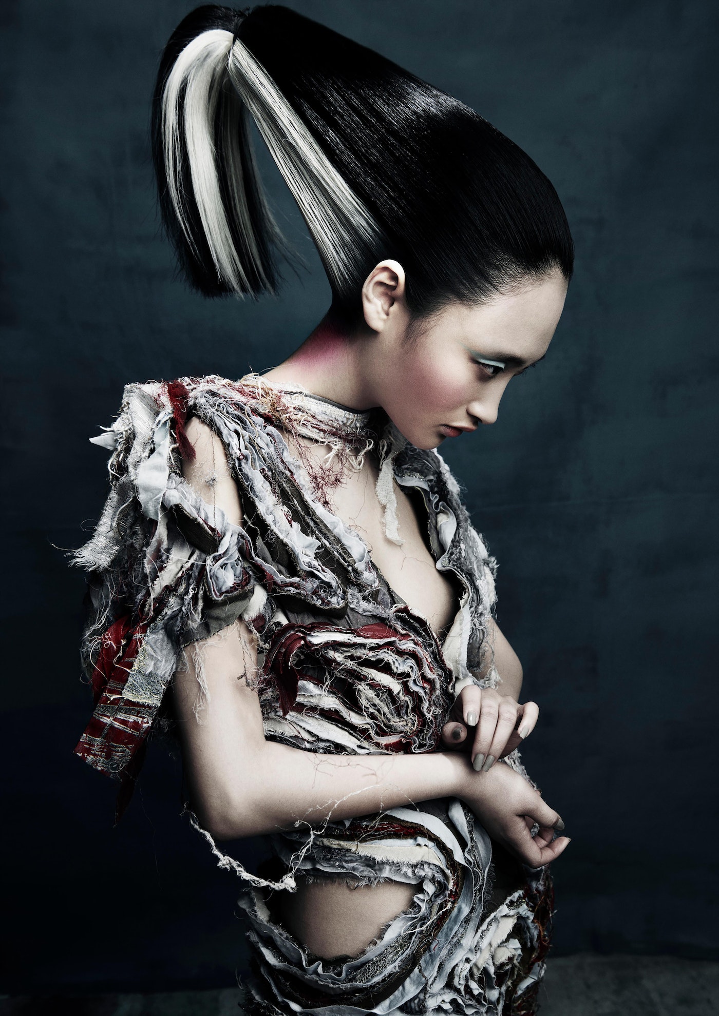 A woman with black and white high ponytail stands with her arm across her torso, wearing tattered white, red, and grey clothing.