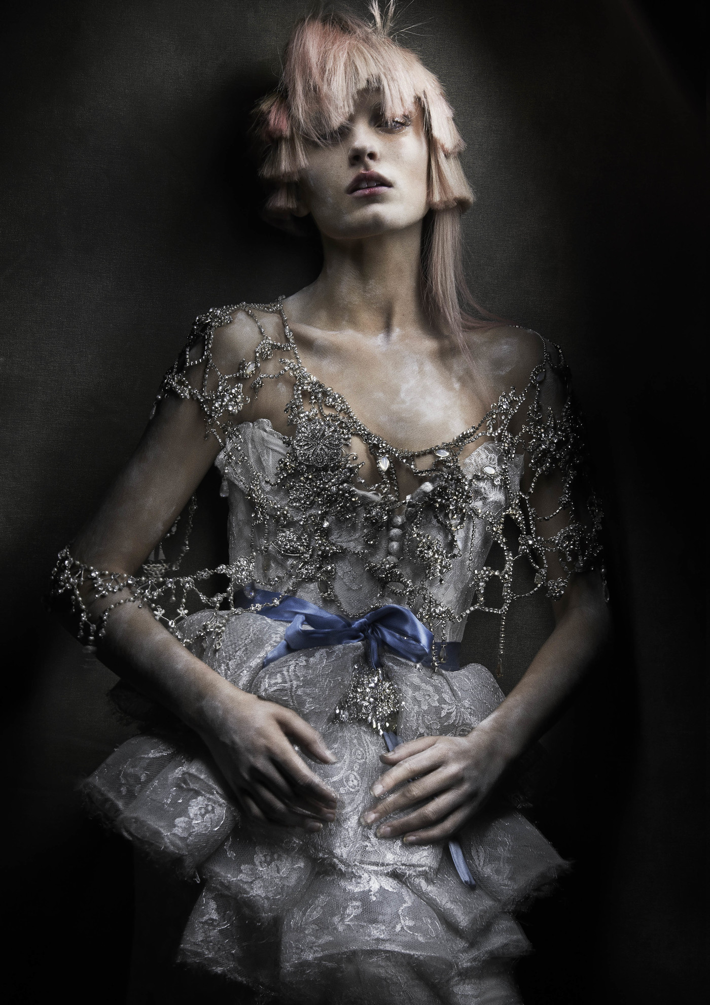 An adult female fashion model poses indoors in a formal dress with lace and beading, featuring stylized pink hair and dramatic makeup.