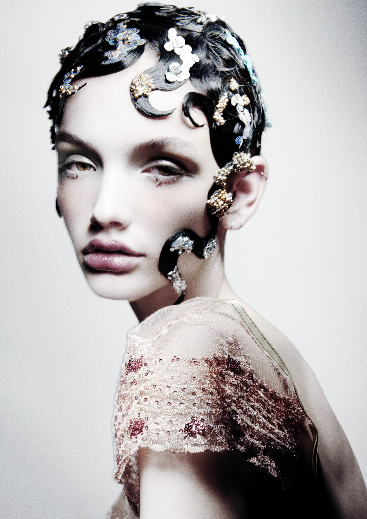 Headshot of a woman with creative styling including black finger-wave hair decorated with white and gold embellishments against a white backdrop.