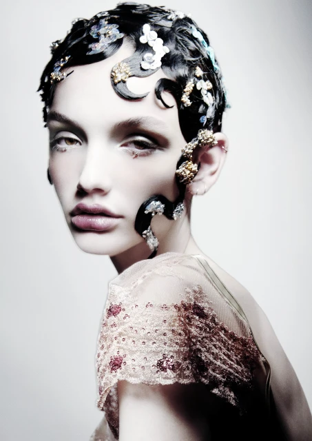Headshot of a woman with creative styling including black finger-wave hair decorated with white and gold embellishments against a white backdrop.
