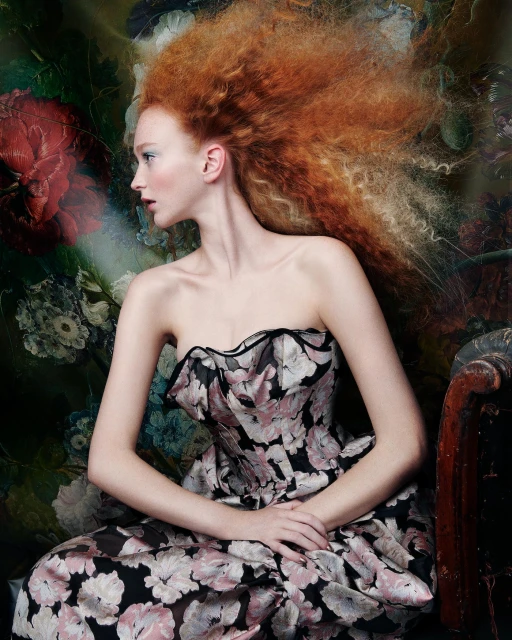 Woman with red hair wearing a floral dress in a studio setting with a painted backdrop and an antique chair.