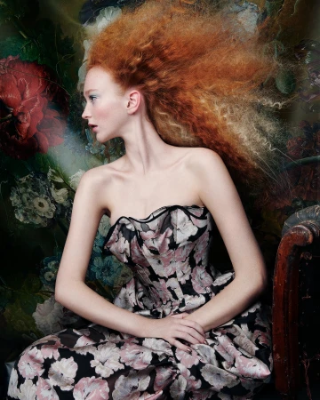 Woman with red hair wearing a floral dress in a studio setting with a painted backdrop and an antique chair.