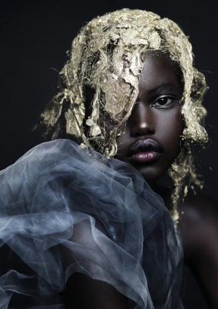 Close-up portrait of a Black woman with gold leaf in her hair and a translucent grey tulle garment by Andrew O’Toole.