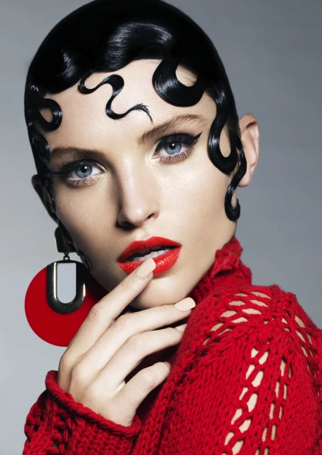 Close-up of a woman with stylized black hair, dramatic winged eyeliner, and bold red lipstick, wearing a red knit sweater and red earrings.